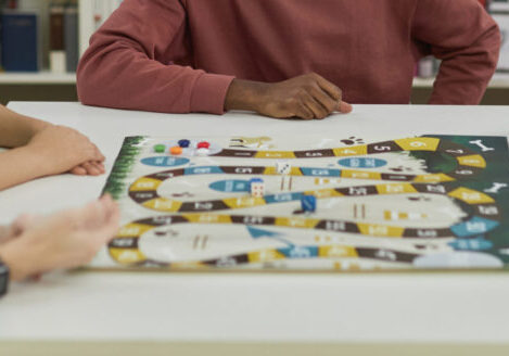 Diverse group of young people playing board games together while sitting around table in library and smiling